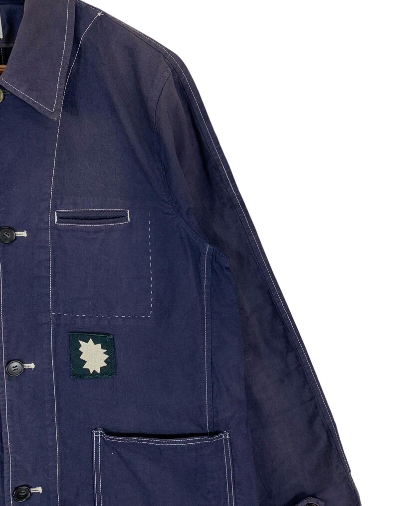 May include: A dark blue long-sleeve jacket featuring a pointed collar, white contrast stitching, and dark buttons. The front includes a chest pocket and a lower pocket, with a small square dark green patch displaying a white star-shaped graphic.