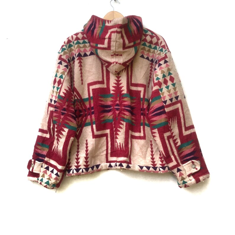 May include: A beige hooded jacket with a geometric pattern in burgundy, green, pink, and purple. The jacket has a button closure and is hanging on a wooden hanger. The design features triangles and cross-like shapes.