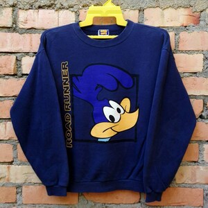 May include: A navy blue sweatshirt featuring a cartoon image of Road Runner from the Looney Tunes cartoon. The image is within a yellow square with the words "Road Runner" in yellow text on the left side of the image.