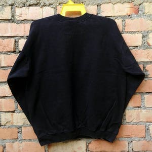 May include: Black sweatshirt with a crew neck and long sleeves. The sweatshirt is hanging on a yellow hanger against a brick wall.