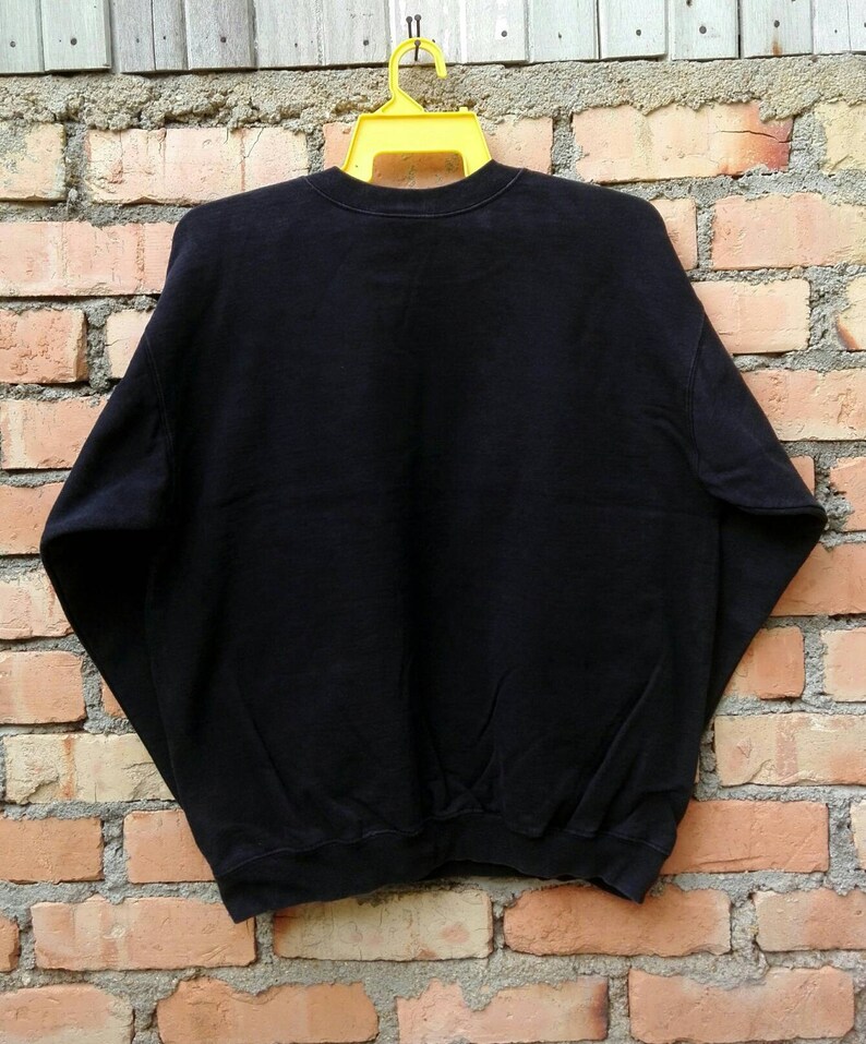 May include: Black sweatshirt with a crew neck and long sleeves. The sweatshirt is hanging on a yellow hanger against a brick wall.