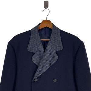 Puede incluir: Un abrigo azul marino de doble botonadura con cuello estampado gris. El abrigo est&aacute; colgado de una percha de madera. El cuello tiene un peque&ntilde;o patr&oacute;n repetitivo. El abrigo tiene dos filas de botones.