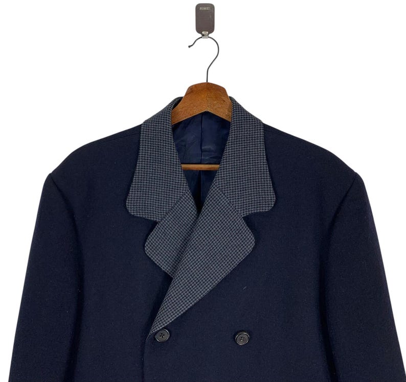 Puede incluir: Un abrigo azul marino de doble botonadura con cuello estampado gris. El abrigo est&aacute; colgado de una percha de madera. El cuello tiene un peque&ntilde;o patr&oacute;n repetitivo. El abrigo tiene dos filas de botones.