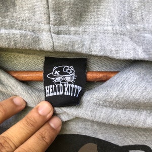 May include: A black and white Hello Kitty label with a cartoon image of Hello Kitty wearing a hat and sunglasses. The label says "Hello Kitty" in white letters.