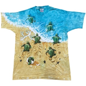 May include: A tie-dye t-shirt featuring a beach scene with sea turtles. The shirt has a blue and white sky and ocean design, with a sandy beach at the bottom. Several green sea turtles are depicted on the beach and in the water.