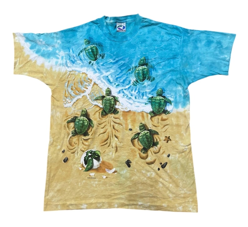 May include: A tie-dye t-shirt featuring a beach scene with sea turtles. The shirt has a blue and white sky and ocean design, with a sandy beach at the bottom. Several green sea turtles are depicted on the beach and in the water.