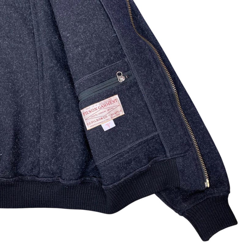 Puede incluir: Chaqueta de lana Filson Garment gris oscuro con cierre de cremallera y bolsillo. La chaqueta tiene dobladillo y pu&ntilde;os acanalados. El bolsillo interior tiene una etiqueta que dice "Filson Garment" y "CC Filson Co. Seattle".