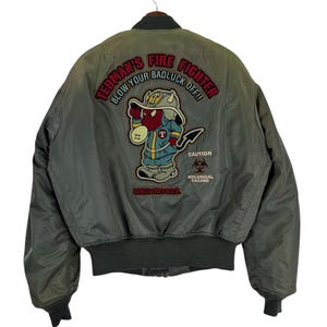 May include: An olive green bomber jacket featuring a large embroidered graphic on the back. The design depicts a red devil character in a firefighter helmet with the text "TEDMAN'S FIRE FIGHTER BLOW YOUR BADLUCK OFF!!" and "CAUTION BIOLOGICAL HAZARD".