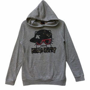 May include: Gray hooded sweatshirt with a Hello Kitty graphic. The graphic features Hello Kitty wearing a black and camouflage baseball cap with a red 'K' on it and red sunglasses. The text 'HELLO KITTY' is printed below the graphic in a black and camouflage font.