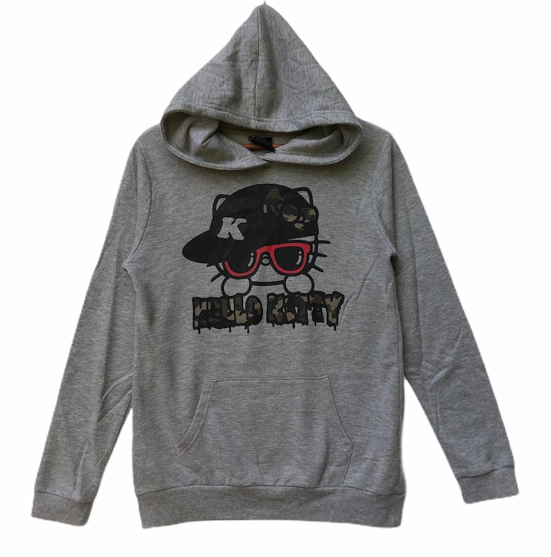 May include: Gray hooded sweatshirt with a Hello Kitty graphic. The graphic features Hello Kitty wearing a black and camouflage baseball cap with a red 'K' on it and red sunglasses. The text 'HELLO KITTY' is printed below the graphic in a black and camouflage font.
