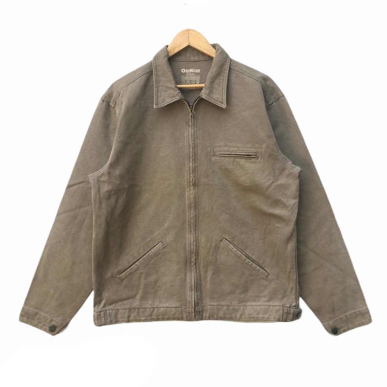 K&ouml;nnte beinhalten: Eine braune Rei&szlig;verschlussjacke mit einem "Orvis"-Etikett. Die Jacke hat zwei Brusttaschen und zwei untere Taschen.