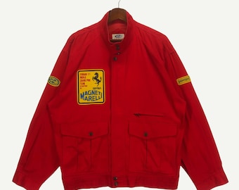 Può includere: Giacca rossa con logo Ferrari e il testo "Ferrari F1 World Champion Grand Prix Team Supported by Magneti Marelli" sul lato sinistro del petto. La giacca ha una chiusura a zip e due tasche.