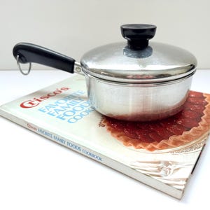 以下が含まれることがあります： 黒いハンドルと蓋が付いた小さなヴィンテージの銀色のソースパン。 「Crisco's Favorite Family Foods Cookbook」というタイトルの料理本の上に置かれています。 料理本の表紙にはパイが描かれています。