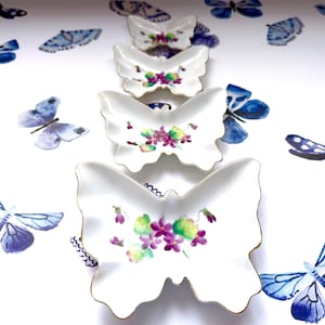 May include: Four white butterfly-shaped dishes with gold trim, arranged in a row. Each dish features a floral design with purple and green accents. The background is white with blue butterfly decals.