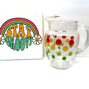 May include: A clear glass pitcher with a handle and a lid, decorated with colorful polka dots. Next to it is a white square with a rainbow and the words "Stay Groovy" in orange and green.