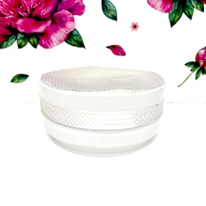 May include: A white cosmetic jar with a textured lid and a decorative band. The jar is set against a white background adorned with watercolor floral elements, including pink peonies and green leaves.