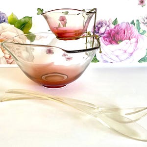 May include: A vintage glass salad bowl set with a smaller bowl suspended above the larger one by a gold-colored metal stand. The bowls have a pink hue and are accompanied by clear plastic serving utensils. Floral background.