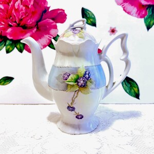 Puede incluir: Una tetera de cerámica blanca con tapa y asa curva. La tetera está decorada con una banda azul y bayas moradas y hojas verdes pintadas. El fondo presenta motivos florales rosas.