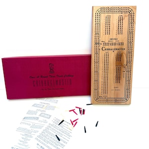 May include: A Drueke Model 1950 Cribbagemaster wooden board with a three-track layout, a red storage box labeled Once-A-Round Three Track Cribbage, a set of black and red plastic pegs, and a printed instruction sheet with diagrams and text.