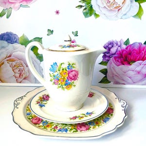 May include: A white porcelain tea set with a floral cross-stitch design. The set includes a teapot, a saucer, and a plate. The teapot and plates have a gold trim. The background features floral wall art.