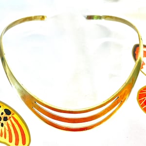 May include: A gold-toned choker necklace with three horizontal cut-out lines. The necklace has a curved design. The background includes colorful butterfly wing designs in orange, yellow, black, and white.