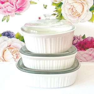 May include: A set of three white ceramic baking dishes stacked vertically, each featuring a vertical ribbed texture on the exterior. The top dish is covered with a clear glass lid that has a small knob handle. The dishes are round in shape.