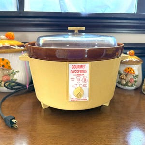 May include: A vintage yellow and brown Gourmet Casserole by American electric appliance with a glass lid. The casserole sits on a wooden surface, with a power cord and plug visible. Decorative mushroom-shaped ceramic jars are in the background.