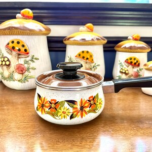 Puede incluir: Un cazo de metal blanco moteado con tapa marrón, mango negro y un vibrante estampado floral naranja y amarillo. Detrás hay varios botes de cerámica con forma de seta, con sombreros marrones y diseños de setas pintados en los cuerpos.