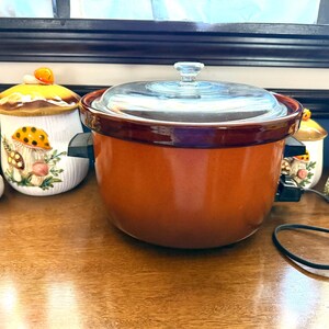 Puede incluir: Una olla de cocción lenta naranja con tapa de cristal y asas negras se encuentra sobre una superficie de madera. Tarros de cerámica decorativos con forma de hongo con detalles amarillos y naranjas están colocados alrededor.
