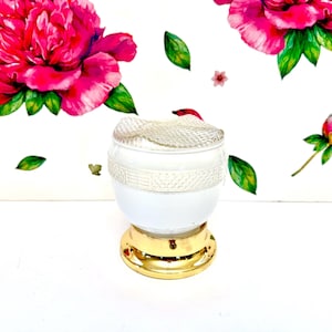 May include: A white and gold decorative jar with a textured glass lid. The jar has a gold base and a white body with a decorative band. The background features floral wall art with pink peonies and green leaves.