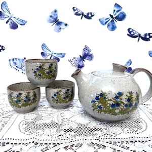 May include: A ceramic tea set with a teapot and three cups, decorated with blue floral patterns and green leaves. The teapot has a brown handle and spout. The background features watercolor butterfly illustrations.