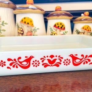 Puede incluir: Una fuente rectangular blanca para hornear con un diseño de pájaros y flores rojas. Detrás de la fuente hay varios botes de cerámica con forma de hongo con detalles amarillos y marrones. Los botes tienen tapas en forma de hongo.