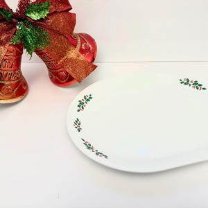 May include: White oval serving platter with green holly and red berry accents. Two red Christmas bells with gold trim and a red bow are in the background. Festive holiday serving dish.