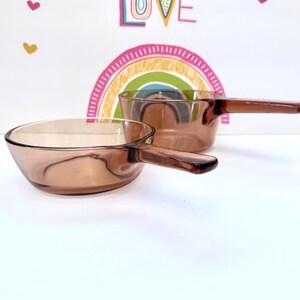 Puede incluir: Dos cacerolas vintage de vidrio ámbar con asas. Las cacerolas son de vidrio transparente con un tono marrón cálido. Las asas también son de color ámbar y parecen estar hechas del mismo material. Las cacerolas están sobre una superficie blanca.