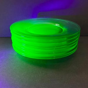 May include: A stack of five circular glass plates that emit a bright, neon green glow when exposed to ultraviolet light. The plates are translucent, featuring a smooth, simple design with a slightly raised center and a flat, circular rim.