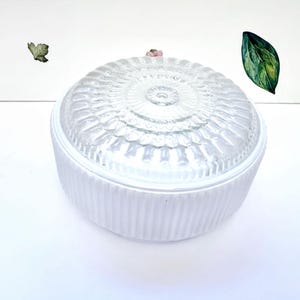Puede incluir: Un recipiente redondo de cerámica blanca con una textura acanalada vertical en los lados y una tapa de vidrio transparente con un patrón radial. La tapa tiene un pequeño pomo central y se asienta sobre la base blanca, formando un cuenco circular.