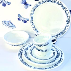 Puede incluir: Un juego de vajilla de cerámica blanca con un diseño floral azul. El juego incluye un plato grande, un cuenco, una taza y un platillo, y platos más pequeños. El fondo presenta una superficie blanca con ilustraciones de mariposas azules.