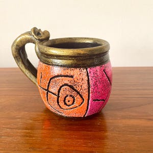 May include: A small, round, orange and pink ceramic mug with a gold rim and a unique, abstract design. The mug has a handle that is shaped like a curved, stylized bird.