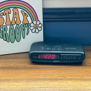 May include: A dark grey Sony Dream Machine clock radio with a digital display showing the time 4:20 in red. The device features an analogue radio tuner dial and control buttons on the top surface. Beside it is a white sign with a rainbow and the text STAY GROOVY.