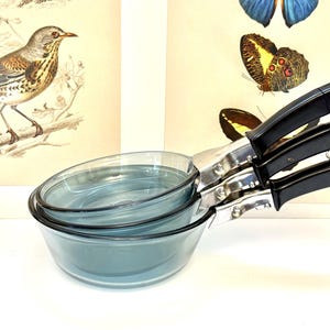 May include: A set of three stacked, round, transparent blue glass saucepans with black plastic handles and silver-toned metal hardware. Each handle features a small metal loop at the end for hanging, and the glass bowls have a smooth, curved design.
