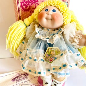May include: A Cabbage Patch Kids doll with yellow yarn hair, blue eyes, and rosy cheeks. The doll wears a light blue dress with white lace trim and blue ribbon accents. The doll is displayed with its original box and certificates.