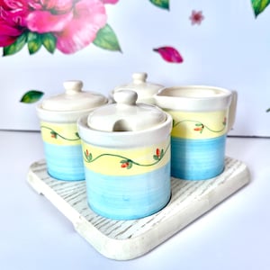 May include: A ceramic serving set with a light blue and yellow color scheme. The set includes two lidded jars, a creamer, and a sugar bowl with a spoon slot, all on a white wooden tray. The jars and creamer are decorated with a floral vine design.