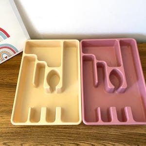 Puede incluir: Dos organizadores de cubiertos de plástico, uno de color amarillo claro y otro de color rosa claro. Ambos organizadores tienen varios compartimentos para separar los diferentes tipos de cubiertos.