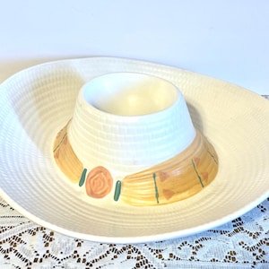 以下が含まれることがあります： 白いセラミック製のソンブレロ型サービングディッシュ。中央にボウルがあり、つばが広い。帽子の帯は、ベージュ、緑、桃色のアクセントで塗装されています。お皿はテクスチャ加工された表面です。