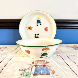 May include: A white ceramic bowl and plate set with a green rim. The bowl and plate are decorated with cartoon figures, hearts, and a sun. The set is on a white tablecloth with a cat illustration.
