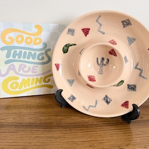 以下が含まれることがあります： 中央にディップボウルが付いた、桃色のセラミックチップ＆ディップサービング皿。皿は赤、青、緑の幾何学模様で装飾されています。「Good Things Are Coming」という言葉が書かれた装飾的なサインも画像にあります。