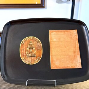 May include: A rectangular black serving tray featuring a circular wooden inlay with the engraved text Schweppes Since 1783 and a fountain graphic, positioned next to a rectangular light brown wooden piece with a horizontal plank pattern.