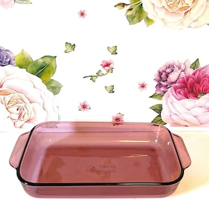 May include: A rectangular, transparent, pink glass baking dish with black trim and handles. The dish is set against a white background decorated with watercolor floral designs, including roses and peonies.