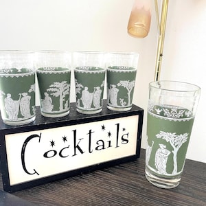May include: Set of five vintage cocktail glasses with a green and white design. The glasses feature a classical scene with figures and trees. The word "Cocktails" is displayed on a sign in front of the glasses.