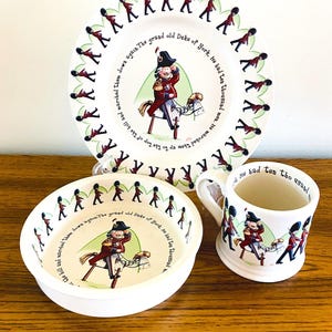 May include: A three-piece ceramic dish set featuring a plate, bowl, and mug. Each piece is decorated with a cartoon illustration of a man on a horse and marching soldiers. The plate and bowl have the text "The grand old Duke of York" and "He had ten thousand men".